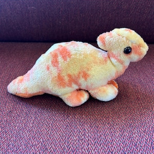 May include: A plush dinosaur toy featuring a soft, fuzzy texture with a tie-dye pattern of light orange and cream colors. The stuffed animal has a rounded head, a small crest, and two black plastic eyes, depicted in a side-facing profile view.