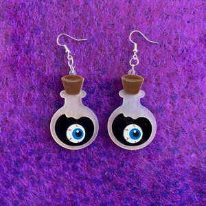 Könnte beinhalten: Ein Paar Halloween-Ohrringe. Jeder Ohrring hat die Form einer durchsichtigen Flasche mit schwarzem Innenraum und einem blauen Augendesign. Sie haben braune Verschlüsse und silberne Ohrhaken. Der Hintergrund ist ein strukturierter Lila-Ton.