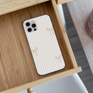 May include: A smartphone case with a cream and beige striped pattern and small bow accents. The case is inside a wooden drawer. The phone case has a camera with three lenses.