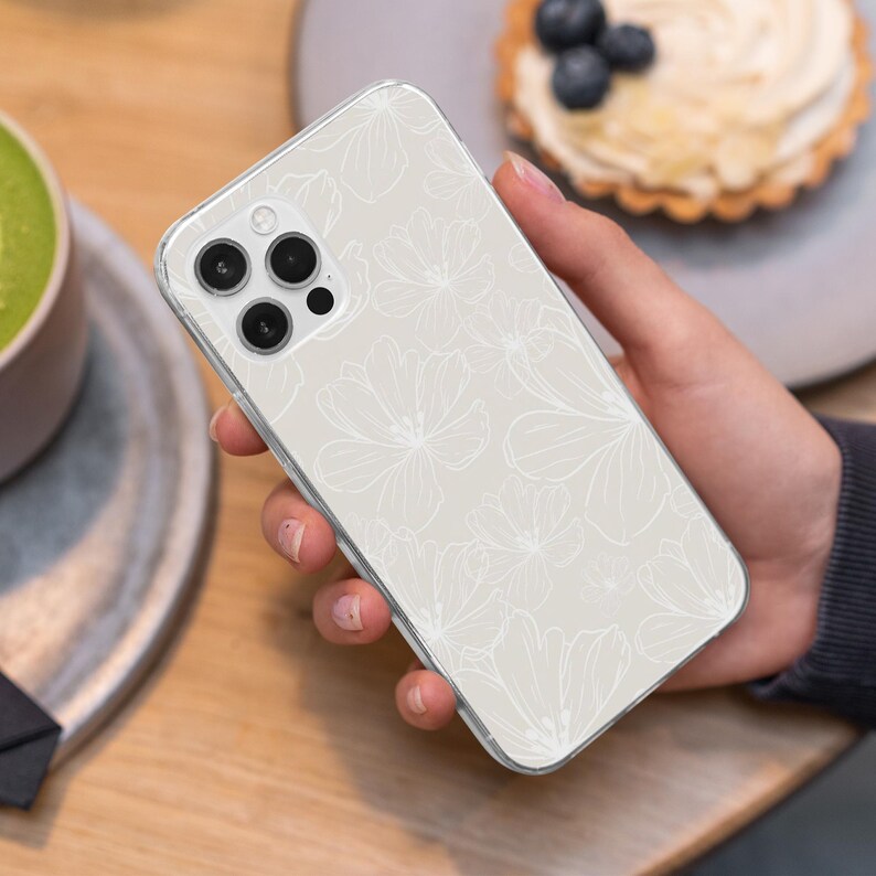 May include: A clear phone case with a floral design. The case has a light beige background with white floral outlines. The phone case is held in a hand, with the camera lenses visible.