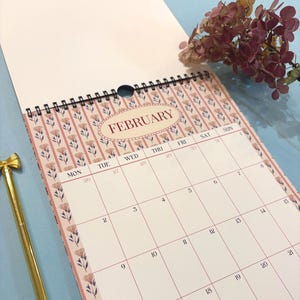 May include: A spiral-bound calendar open to February. The calendar features a floral design in pink and cream, with the month's name in a decorative oval. A gold pen with a bow-shaped top is in the lower left corner.