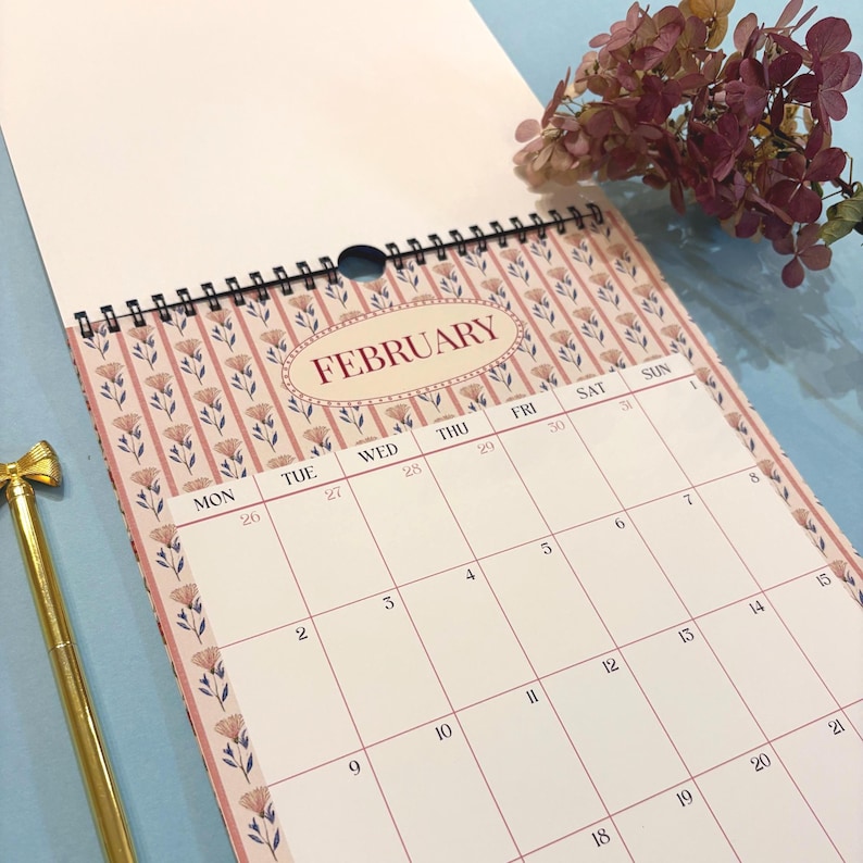 May include: A spiral-bound calendar open to February. The calendar features a floral design in pink and cream, with the month's name in a decorative oval. A gold pen with a bow-shaped top is in the lower left corner.