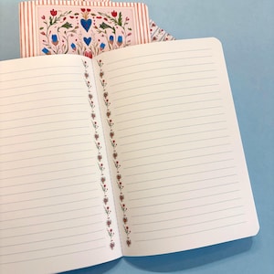 May include: An open notebook with lined pages and a floral border. The cover displays a floral design with blue hearts and red flowers on a striped background. The notebook is on a blue surface.