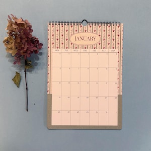 May include: A wall calendar for January with a floral design and a gray bottom. The calendar has a spiral binding and a black loop for hanging. A dried flower arrangement is to the left of the calendar.