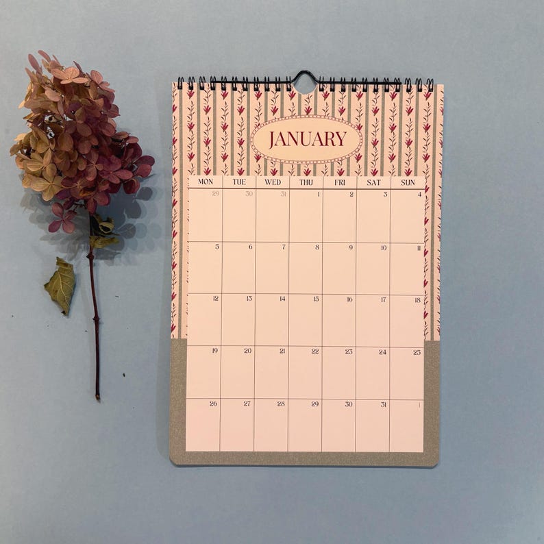 May include: A wall calendar for January with a floral design and a gray bottom. The calendar has a spiral binding and a black loop for hanging. A dried flower arrangement is to the left of the calendar.