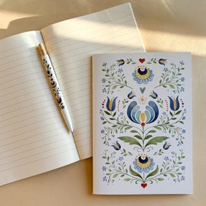 Könnte beinhalten: Ein weißes Notizbuch mit linierten Seiten und ein passender weißer Stift, beide verziert mit einem symmetrischen Blumenmuster aus blau, gelb und grün gefärbten Aquarellblumen, Blättern und kleinen roten Herzen auf weißem Grund.