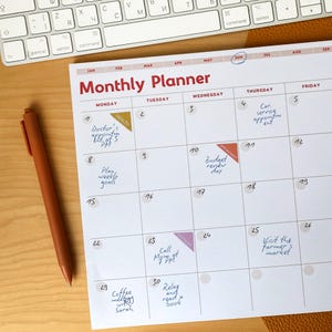 Puede incluir: Un planificador mensual con el título "Monthly Planner" en rojo. La cuadrícula del calendario está llena de notas manuscritas y pegatinas. Un bolígrafo naranja descansa sobre la superficie de madera junto al planificador y un teclado blanco.