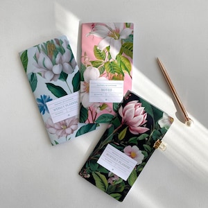 Op de afbeelding: Drie notitieboekjes met bloemmotieven en verschillende titels: "Notities", "Wekelijkse Planner" en "Maandelijkse Planner". Elk notitieboekje heeft een wit label met de tekst "Esthetisch en Functioneel" en "Botanisch" erop gedrukt.