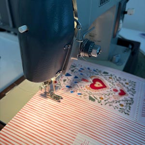 May include: A close-up of a vintage sewing machine in operation, stitching a planner with a red and white striped pattern and a floral design with red hearts. The machine is grey and blue, with the needle and thread in focus.