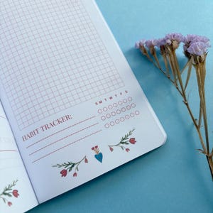 May include: Open notebook with a habit tracker grid and the text "HABIT TRACKER:". The pages have a pink grid pattern and floral illustrations. A sprig of dried purple flowers rests on a blue surface.