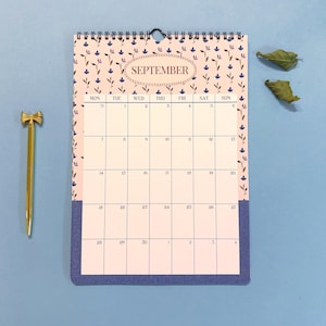 May include: A spiral-bound wall calendar for September with a floral pattern and the month's name in an oval. A gold pen with a bow-shaped top rests beside the calendar on a blue surface.