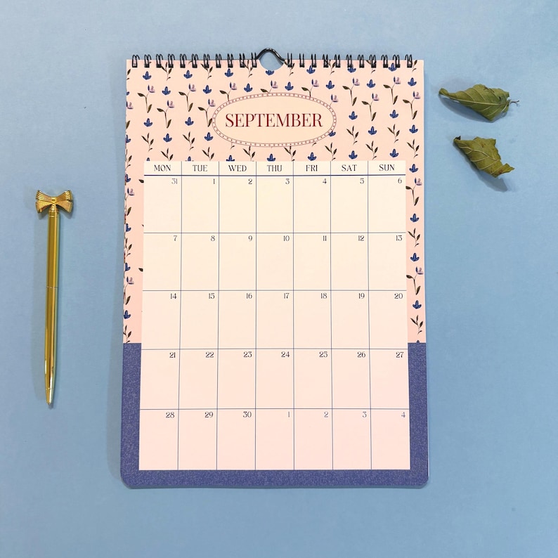 May include: A spiral-bound wall calendar for September with a floral pattern and the month's name in an oval. A gold pen with a bow-shaped top rests beside the calendar on a blue surface.
