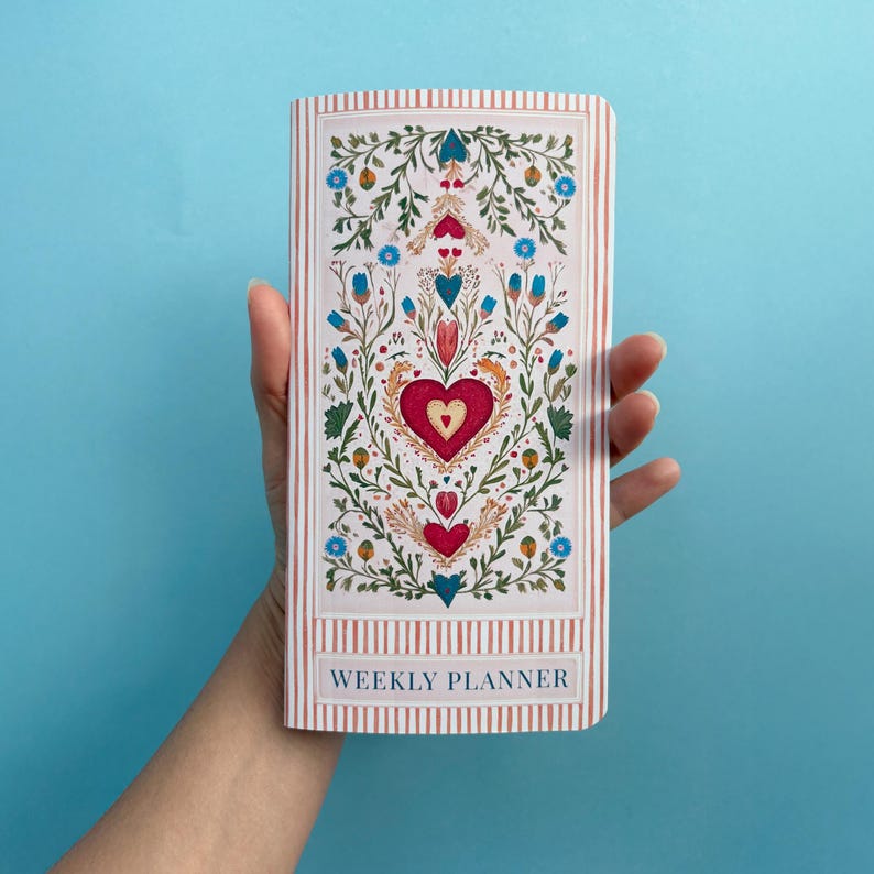 May include: A weekly planner with a floral and heart design. The cover features a red heart at the center, surrounded by green leaves, blue and orange flowers, and smaller hearts. The background has vertical pink and white stripes, with the words "WEEKLY PLANNER" at the bottom.