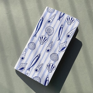 Könnte beinhalten: Ein gefaltetes weißes Tuch mit blauem Streifenmuster. Das Tuch zeigt blaue Illustrationen von Muscheln und Fischen. Das Tuch liegt auf einer grauen Oberfläche.