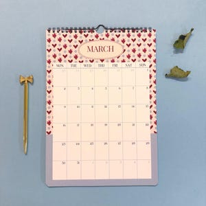 May include: A wall calendar for March with a heart pattern border and the word "MARCH" in an oval. A gold-colored pen with a bow-shaped top is next to the calendar. The calendar is on a blue surface.