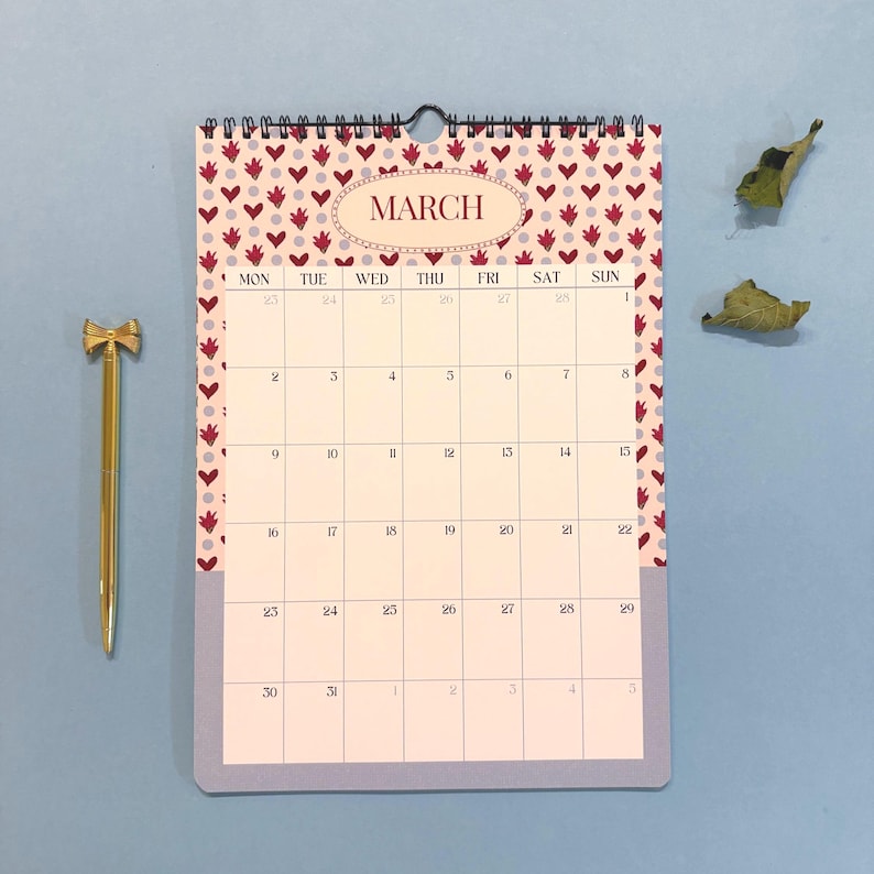 May include: A wall calendar for March with a heart pattern border and the word "MARCH" in an oval. A gold-colored pen with a bow-shaped top is next to the calendar. The calendar is on a blue surface.