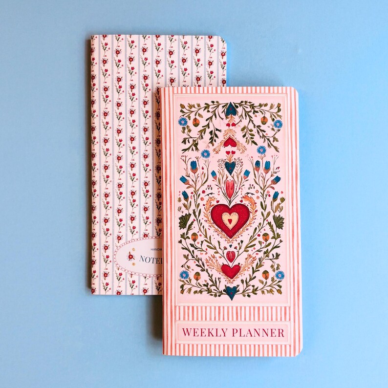 May include: Two notebooks. One has a floral pattern of red flowers on a white background with vertical stripes. The other has a pink background with a heart and floral design and the words "WEEKLY PLANNER".