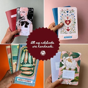 May include: A collection of handmade notebooks with various covers. One red notebook has "NOTES" on a label. Other notebooks feature illustrations of a fashion portrait, floral designs, and fish. The text "WEEKLY PLANNER" is visible on some.