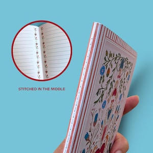 May include: A floral patterned notebook with a cream background and red, blue, and green accents. The notebook is stitched in the middle and has lined pages. The cover features a detailed floral design. The text "STITCHED IN THE MIDDLE" is visible.