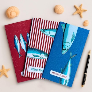 May include: Three colourful planners with fish illustrations. The red planner has a white and red striped design and says "Weekly Plan". The blue planner has a white and blue striped design and says "Monthly Planner".