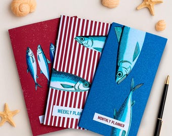 May include: Three colourful planners with fish illustrations. The red planner has a white and red striped design and says "Weekly Plan". The blue planner has a white and blue striped design and says "Monthly Planner".