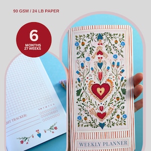 May include: A weekly planner featuring a floral heart design and the words "WEEKLY PLANNER." The planner has a red and white striped border and a habit tracker. The text "90 GSM / 24 LB PAPER" is visible.
