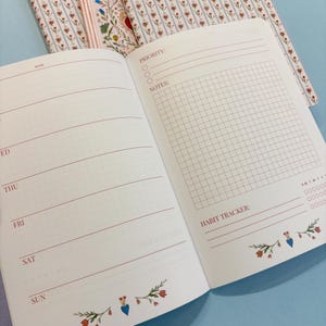 May include: Open notebook with a floral patterned cover. The interior pages are white with red lines for daily planning, a habit tracker, and a grid for notes. Floral and heart designs decorate the bottom of the pages.