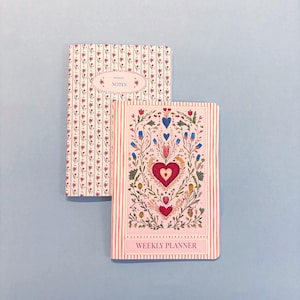 May include: Two notebooks with floral and heart designs on a light blue background. One notebook has a floral pattern and the word "NOTES". The other notebook is a weekly planner with a heart and floral design and the words "WEEKLY PLANNER".