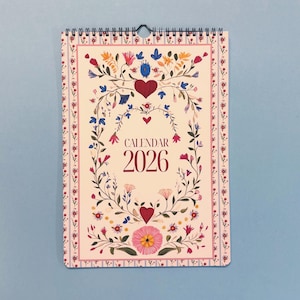 May include: A floral 2026 calendar with a spiral binding. The calendar features a cream background with a decorative border and a central design of flowers, leaves, and hearts. The word "CALENDAR" and the year "2026" are printed in a burgundy color.