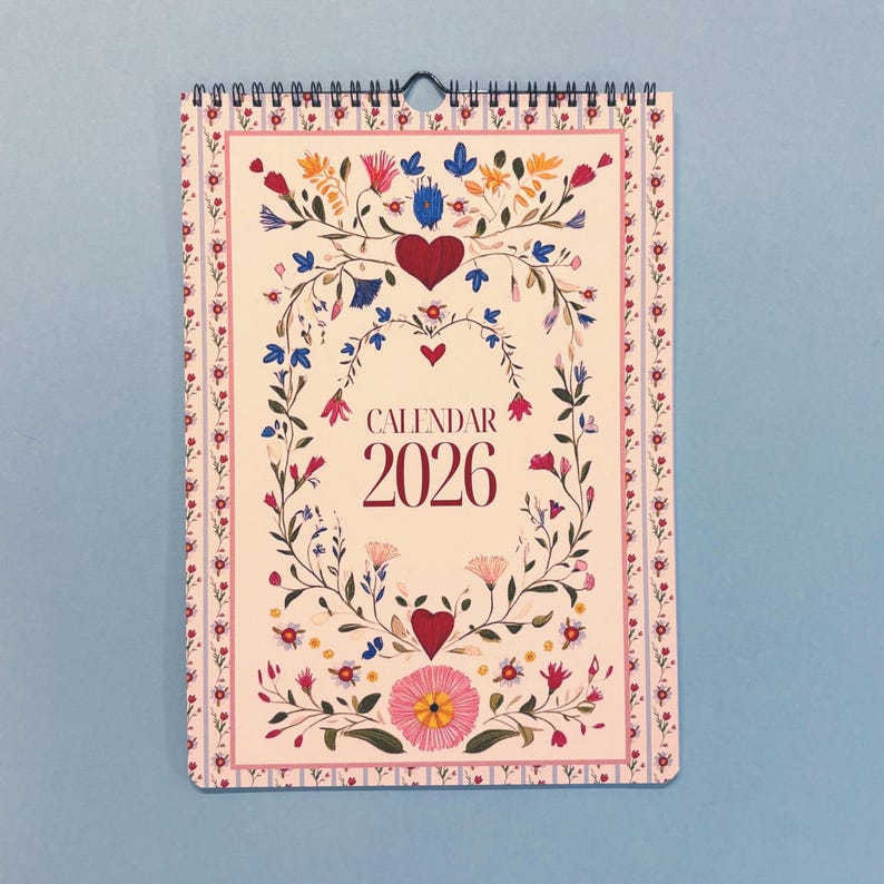 May include: A floral 2026 calendar with a spiral binding. The calendar features a cream background with a decorative border and a central design of flowers, leaves, and hearts. The word "CALENDAR" and the year "2026" are printed in a burgundy color.