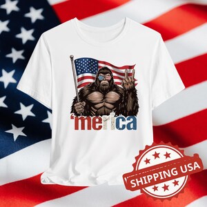 Può includere: T-shirt bianca con un disegno di Bigfoot con occhiali da sole con un design a bandiera americana. Il Bigfoot tiene in mano una bandiera e fa il segno della pace. La parola "merica" è stampata in rosso e blu.