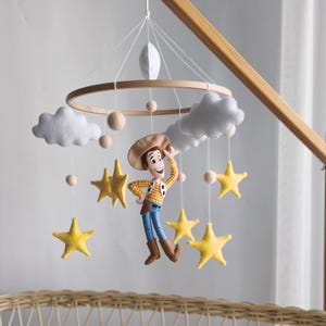Può includere: Mobile per bambini in legno con il personaggio di Woody, stelle gialle, nuvole bianche e perline di legno. Il mobile è appeso sopra una culla in vimini con una coperta bianca.
