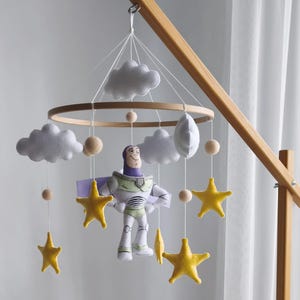 Può includere: Mobile per bambini in legno con un personaggio di Buzz Lightyear, stelle gialle, nuvole bianche e perline di legno. Il mobile è appeso sopra una culla in vimini con una coperta bianca. Il mobile è sostenuto da un telaio in legno.