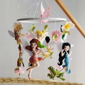 Könnte beinhalten: Ein Babymobile mit Filzfeen, Blumen und einem Frosch, aufgehängt an einem Holzrahmen. Das Mobile hängt über einer Weidenkorb-Wiege mit einer weißen Decke. Rosa und weiße Akzente verleihen der Kinderzimmerdekoration einen verspielten Touch.