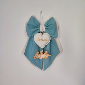 Può includere: Un fiocco azzurro chiaro con un'etichetta bianca a forma di cuore su cui è scritto "Lorenzo". Un piccolo cucciolo di leone di peluche è appeso alla parte inferiore del fiocco.