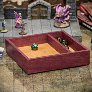 Puede incluir: Una bandeja de dados de madera color burdeos con dos compartimentos, uno forrado con fieltro beige. Un dado verde y dorado descansa en la sección forrada de fieltro. Otros dados y figuras en miniatura están en el fondo, lo que sugiere un tema de juego de mesa.