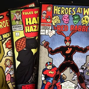 May include: A stack of vintage comic books featuring colorful superhero illustrations. The covers display titles like "Heroes at Work" with "The Red Rabbit" and "Tales of Haphazard!" with "Earth's Mountain". The comics are from Berkshire Comics Group.