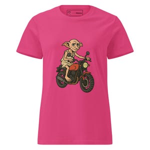 Puede incluir: Camiseta rosa intenso con una ilustración de dibujos animados de un personaje montando en moto. El personaje tiene orejas grandes y viste una túnica clara. La moto es naranja y negra.