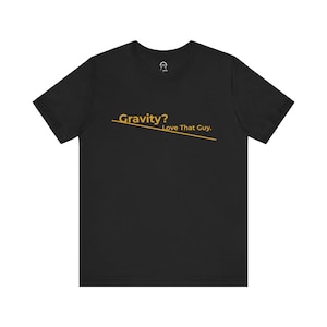 以下が含まれることがあります： 「Gravity? Love That Guy.」という金色のグラフィックがプリントされた黒いTシャツ