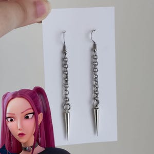 May include: A pair of silver-toned earrings with a chain design and a pointed, cone-shaped pendant. The earrings are displayed on a white card, held by a hand. The image also features a cartoon character.