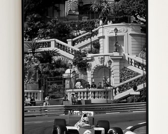 May include: A black and white photograph of a race car driving on a track in Monaco. The car is a Formula One car with the Marlboro logo on the side. The car is driving past a building with a staircase.