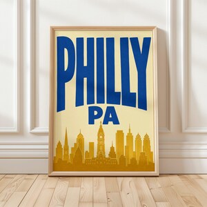 Può includere: Una stampa artistica incorniciata con sfondo color crema e il testo blu in grassetto PHILLY PA nella parte superiore. Sotto il testo si trova una silhouette giallo oro dello skyline della città di Filadelfia, inclusa la torre del municipio.