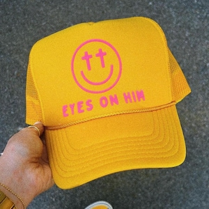 May include: A bright yellow trucker hat featuring a pink graphic of a smiley face with cross-shaped eyes. Below the smiley face, the text EYES ON HIM is printed in pink. The hat has a structured front panel and a mesh back for ventilation.