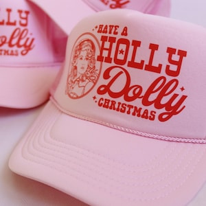 May include: A group of light pink trucker hats featuring a red graphic print on the front panel. The design includes a portrait of a woman inside an oval frame, accompanied by the text "HAVE A HOLLY DOLLY CHRISTMAS" in a bold, stylized font.