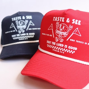 May include: A navy blue and a bright red trucker hat, each featuring a white rope detail across the brim. The front panels display a white graphic of a cartoon burger with the text TASTE & SEE THAT THE LORD IS GOOD PSALM 34:8.