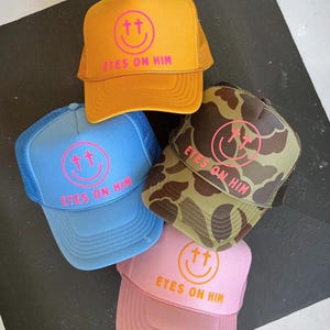 May include: Four trucker hats in yellow, light blue, camouflage, and pink. Each hat features a front graphic of a pink or orange smiley face with cross-shaped eyes and the text EYES ON HIM printed in bold capital letters below the smiley face.