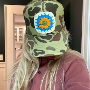 Puede incluir: Gorra de camionero de camuflaje con un parche cuadrado de ganchillo. El parche presenta un borde blanco, un diseño floral azul y naranja y un centro verde. La gorra tiene una parte trasera de malla y una visera curva.