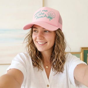 Woman modeling light pink trucker hat with teal puff vinyl design that says "meet me at the beach" in script font beach vacation hat