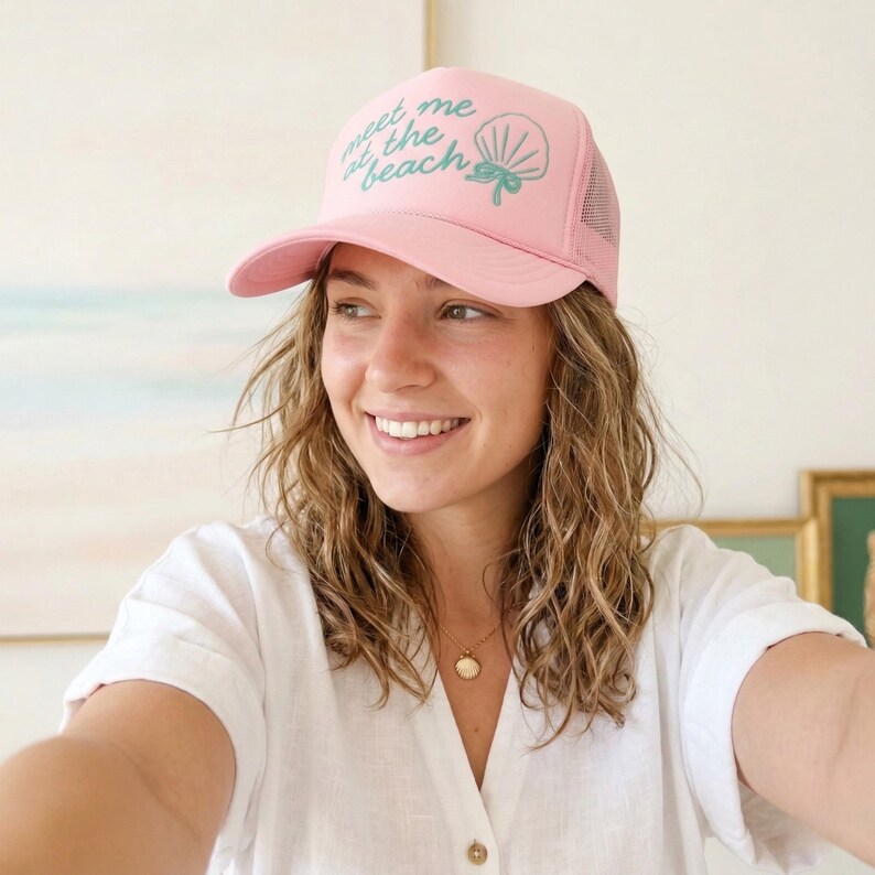 Woman modeling light pink trucker hat with teal puff vinyl design that says "meet me at the beach" in script font beach vacation hat