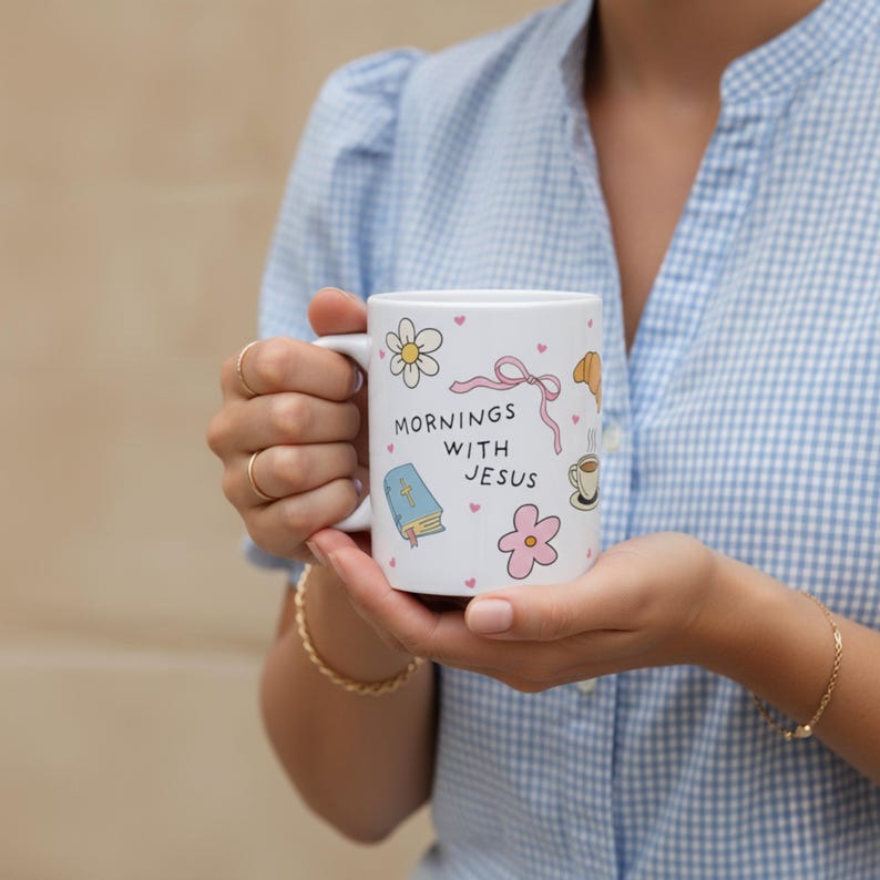 Woman holding white Christian coffee mug with “Mornings with Jesus” text, pastel doodle Bible, flowers, bows, and pink hearts, faith gift idea.